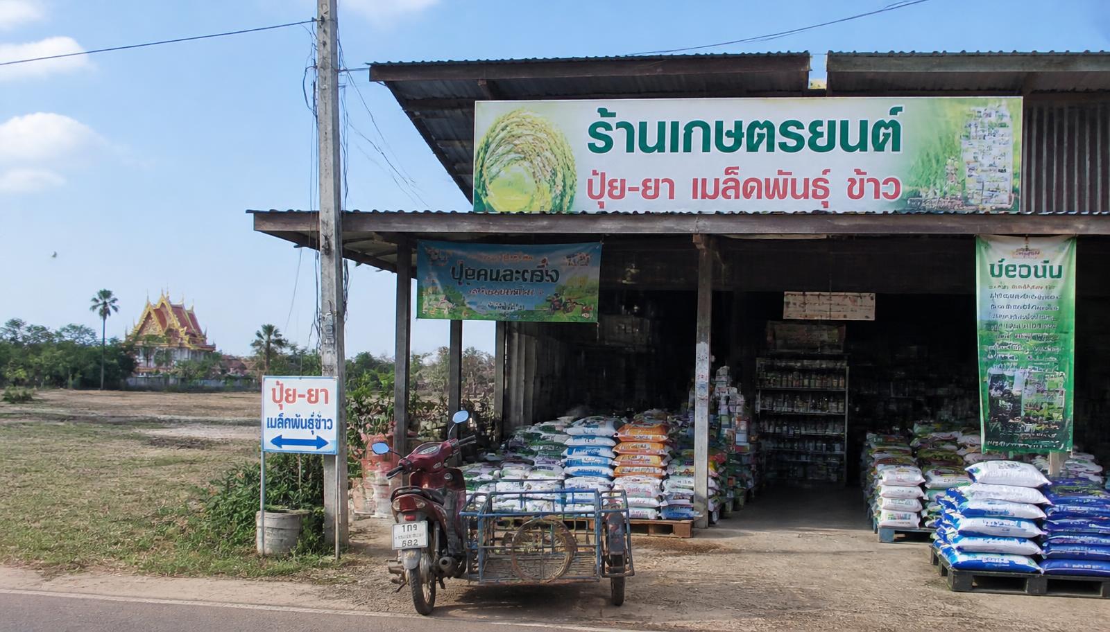 ร้านขายปุ๋ย ยา และเมล็ดพันธุ์ข้าวริมถนนในอำนาจเจริญ มีกระสอบวางเรียงหน้าร้าน รถพ่วงข้าง และวัดอยู่ไกลออกไป