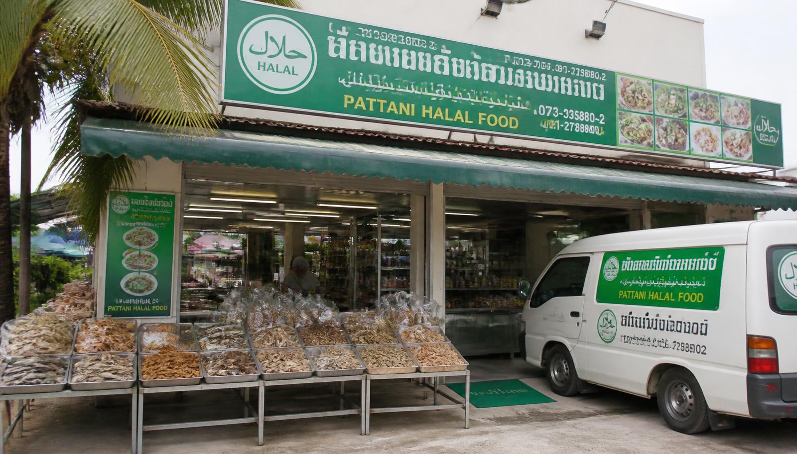 ร้านอาหารฮาลาลในปัตตานี มีถาดปลาแห้งและของแห้งวางหน้าร้าน ป้ายภาษาไทยและอาหรับ และรถตู้ส่งของจอดอยู่
