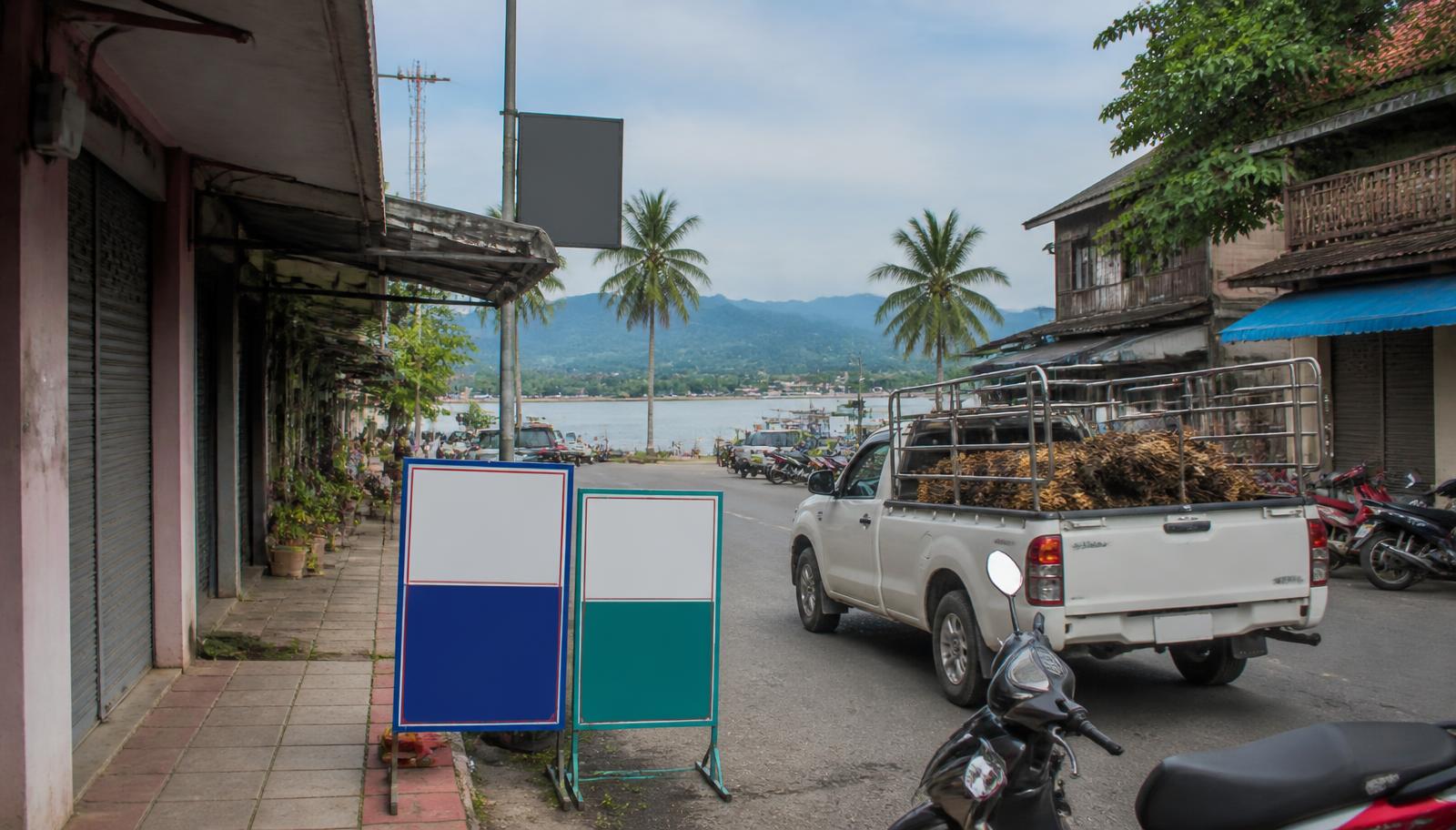 ถนนย่านการค้าใกล้ท่าเรือในจังหวัดสุราษฎร์ธานี มีอาคารพาณิชย์เก่า รถกระบะบรรทุกผลปาล์ม และแผงขายของ