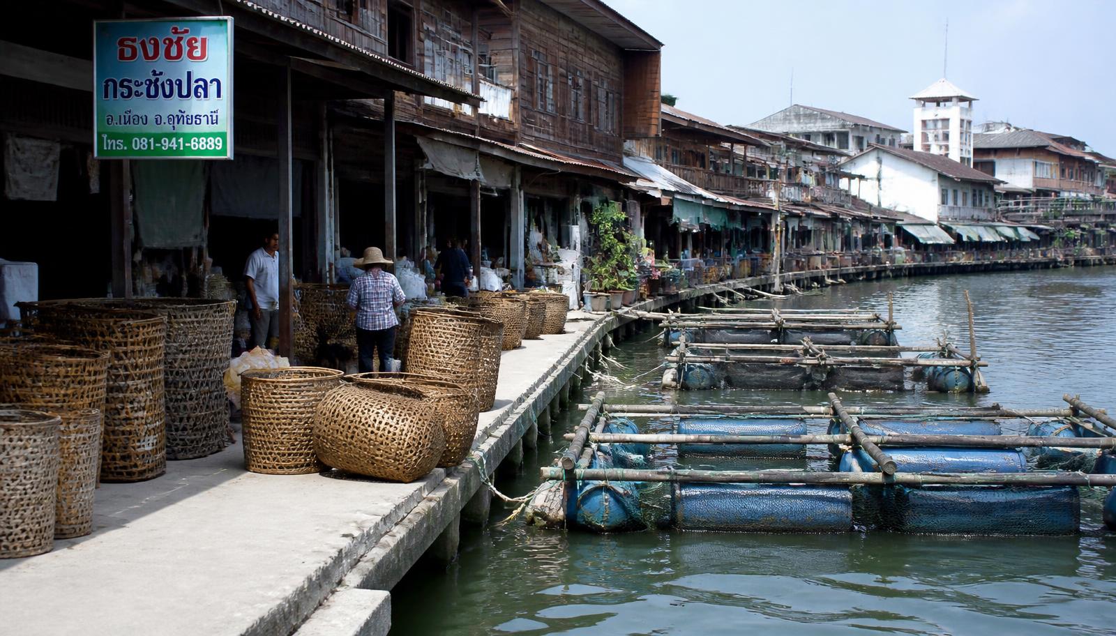 ตลาดริมแม่น้ำสะแกกรังในอุทัยธานี มีกระชังปลาลอยน้ำ เรือนไม้เก่า และตะกร้าไผ่วางเรียงบนทางเดิน